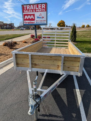 A Silver Ox single-axle aluminum utility trailer with a wooden side, displayed at a dealership lot with a for-sale sign in the background.