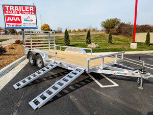 A new Silver Ox Aluminum tandem axle aluminum utility trailer with side ramps displayed at a dealership lot, with a for sale sign in the background.