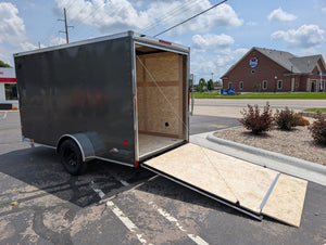 American Hauler enclosed trailer with a wooden ramp on a paved surface under a cloudy sky.